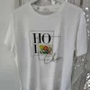 Camiseta blanca con letras en cursiva, flores bordadas en relieve y tachuelas metalizadas en el delantero